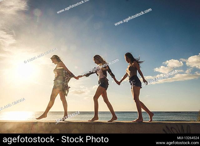 Three young women friends walk together in group enjoying the sunset on the coast - concept of happy young people girls in friendship enjoying the outdoor...