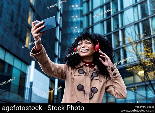 Happy young woman wearing wireless headphones taking selfie through mobile phone in city