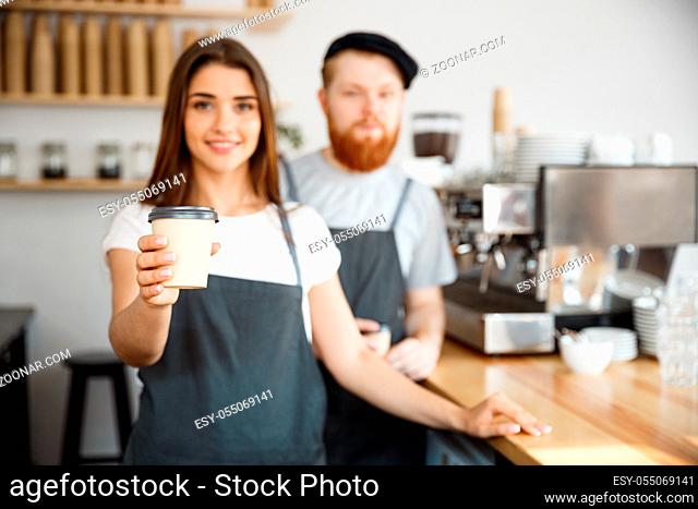 Coffee Business Concept - Positive young bearded man and beautiful attractive lady barista couple giving take away cup of coffee to custome at the modern coffee...