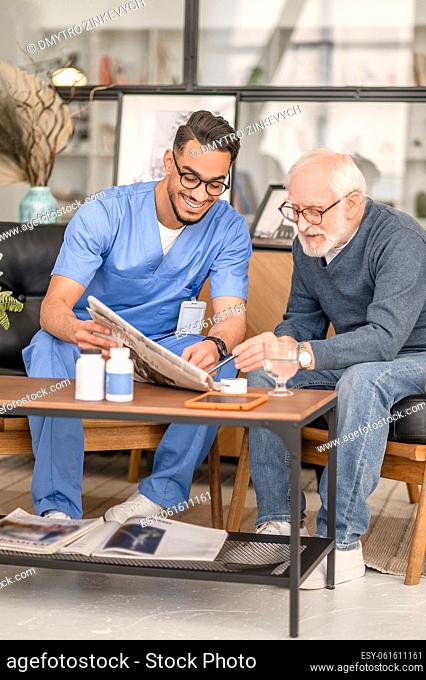 Smiling caregiver and a concentrated aged person sitting at the coffee table and perusing a newspaper article