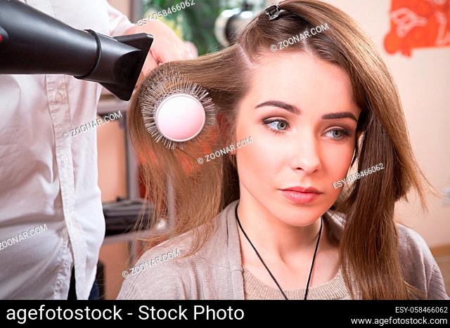 Beautiful lady having her hair dry in hairdressing saloon. Pretty lady looking at mirror while professional hair stylist working