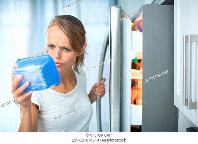 Is this still fine? Pretty, young woman in her kitchen by the fridge, looking at the expiry date of a product she took from her fridge -