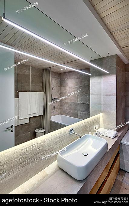 Contemporary bathroom with textured tiles and a wooden ceiling. There is a white sink on the rack, faucet, wooden lockers, mirror, towel, bath, door
