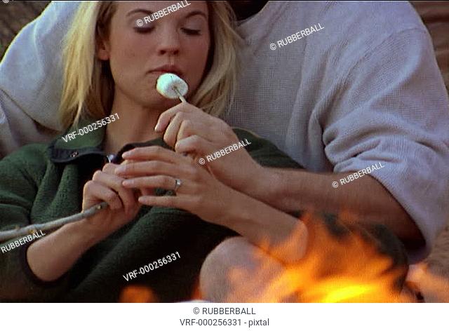 medium two shot os a young caucasian couple as they sit at a camp fire and playfully flirt while roasting marshmallows