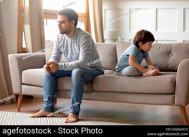Full length annoyed young father sitting back to back with little son on sofa, ignoring each other after argue. Disappointed by bad kids behavior dad not...