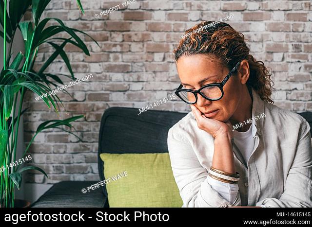 Thoughtful sad young woman in casual clothes sitting on couch in cozy modern living room looking down at home. Depressed caucasian female in deep thoughts...