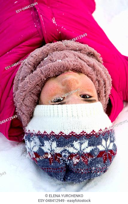 Portrait of a teenage girl in a bright pink winter jacket and knitted hat in the winter in the open air, she lies on pure white snow