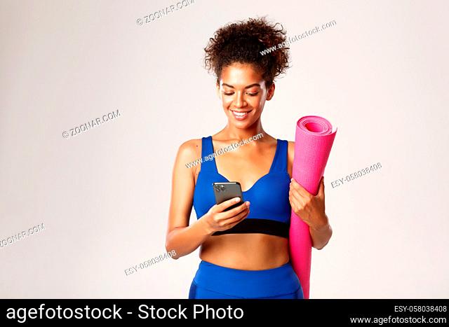 Young athletic woman in blue workout clothing, holding rubber mat for yoga and using mobile phone, standing against white background