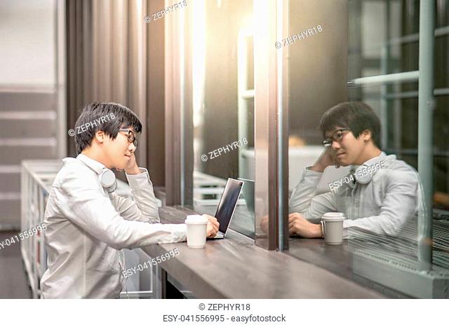 Reflection of young Asian man dressed in casual style working with his laptop computer in cafe. Digital nomad and IT modern lifestyle