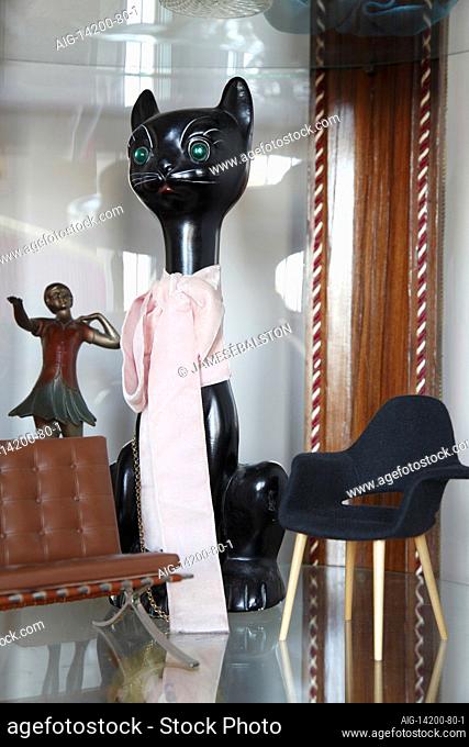 Ceramic black cat with pink bow, 1920s dancer, and miniature modernist chairs in display cabinet | NONE |
