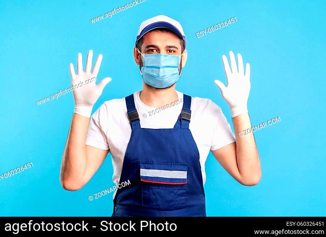 Cheerful handyman in overalls and mask raising hands with surgical gloves, smiling to camera. Safety clothes, protection and hygiene in service industry