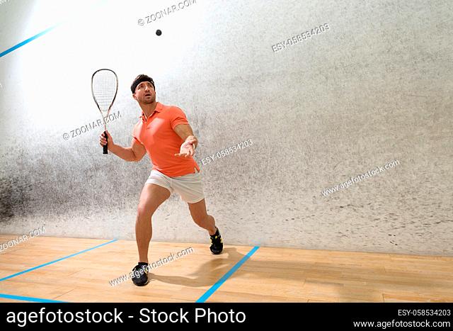 Young squash player hiting ball in squash court. Handsome man in orange t-shirt and white shorts playing popular game in squash