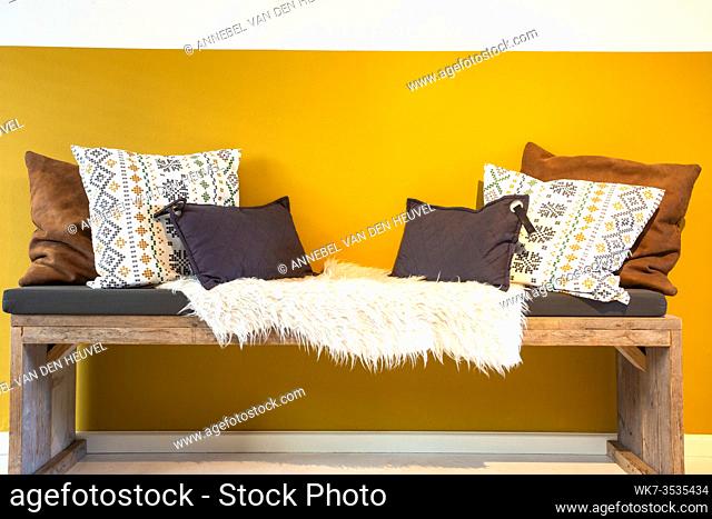 Retro modern wooden bench with white, brown and beige pillows near bright yellow wall Interior of modern home. cozy spot closeup