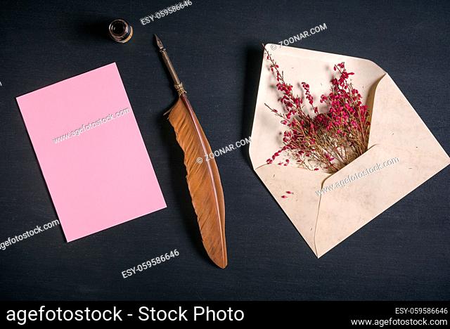 Vintage envelope full with pink flowers, an antique quill pen, an ink pot and a blank message card, on a black wooden background