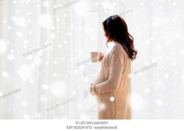 pregnancy, drinks, winter, people and expectation concept - close up of happy pregnant woman with cup drinking tea looking through window at home over snow