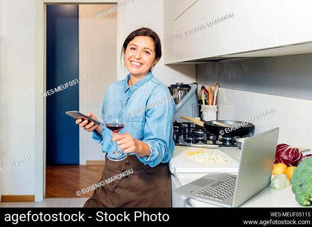 Woman holding wineglass and smart phone leaning on kitchen counter at home