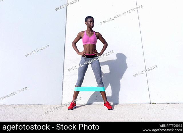 Fit young woman with rubber band doing leg exercise while standing against white wall