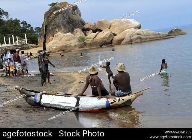 Two people are seen sitting on a canoe as children play in the water while others walk on the beach on the shores of Lake Malawi. Salima, Malawi