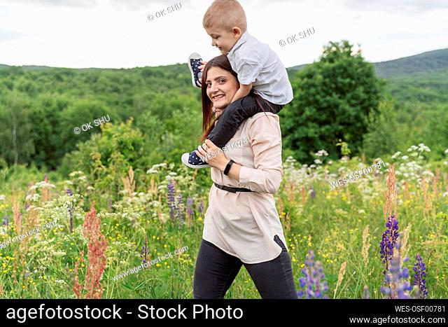 Smiling young woman carrying son on shoulders in meadow