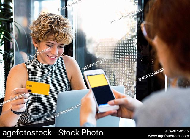 Smiling businesswoman with credit card by colleague using smart phone in cafe