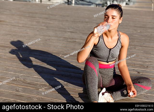 Close-up of attractive sportswoman taking a break after workout, sitting at the seaside and drinking water, looking pleased