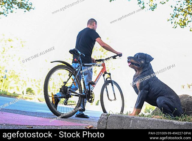 PRODUCTION - 06 October 2023, Baden-Württemberg, Esslingen am Neckar: A barking plastic dog stands on a sidewalk while a cyclist pushes his bike on the sidewalk...