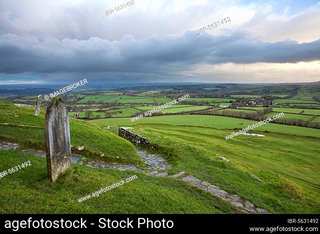 View of graveyard and path leading from 13th century church onto moorland at sunrise, with rain clouds over distant landscape, St Michael's Church, Brent Tor