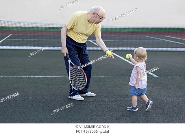 Girl giving tennis ball to grandfather while standing at playing field