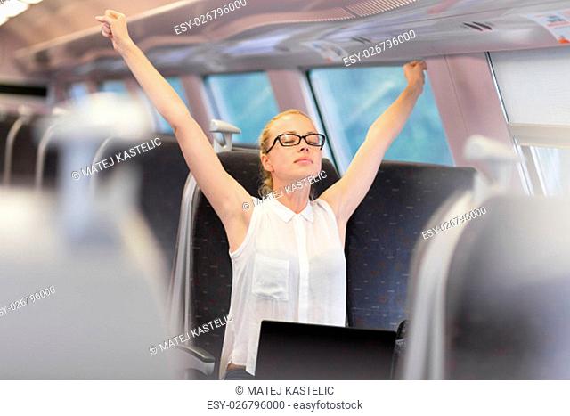 Businesswoman traveling by train, streching her upper body arms reised taking a break from laptop work. Business travel concept