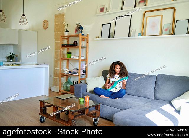 Afro young woman playing ukulele while sitting on sofa at home