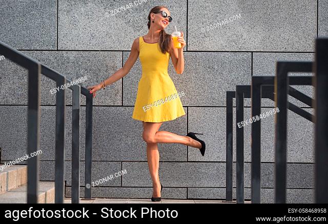 Fashion photo of young magnificent woman in sunglasses. Long-haired girl in little yellow dress posing with orange cocktail