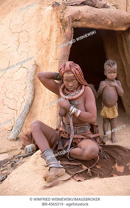 Himba woman with child in front of the mud hut, rubs itself with Okra, butterfat with ochre paint, Kaokoveld, Namibia