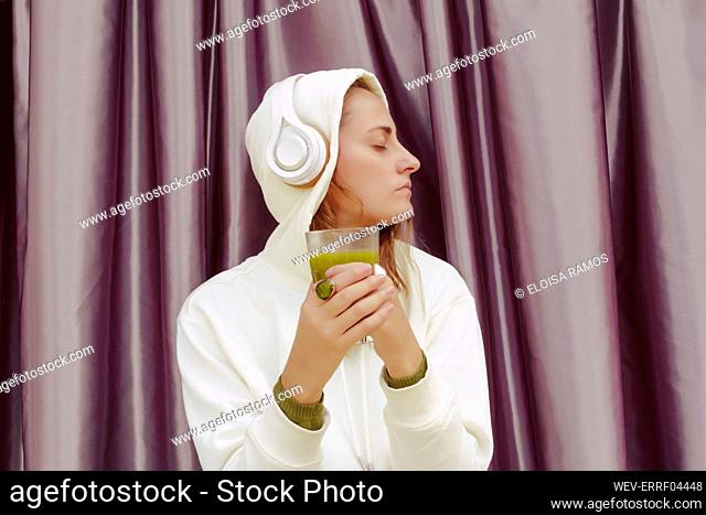 Woman with green juice listening music over headphones while standing against curtain at home