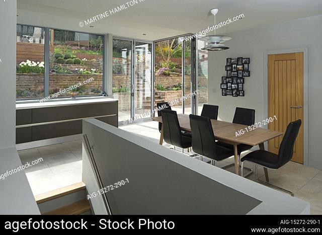 A large open plan area, a kitchen and dining area, with views out of the large full size glass wall panels onto a patio. Minimalist clean lines and plain walls