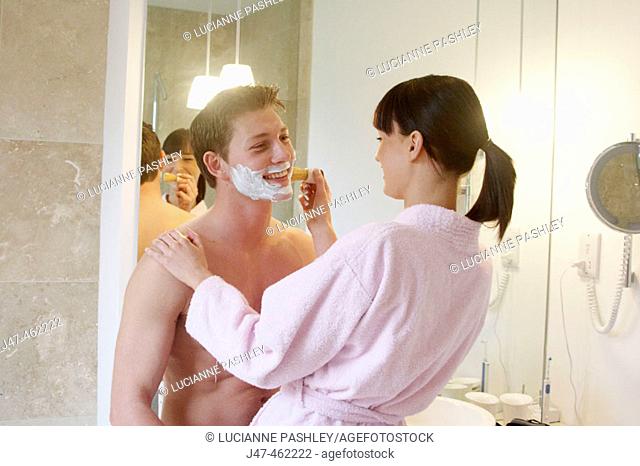 Young couple standing together in underwear in the bathroom, she is helping him shave, both smiling