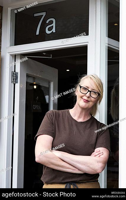 Portrait of waitress with blond hair and glasses, wearing brown apron, leaning against entrance door, smiling at camera