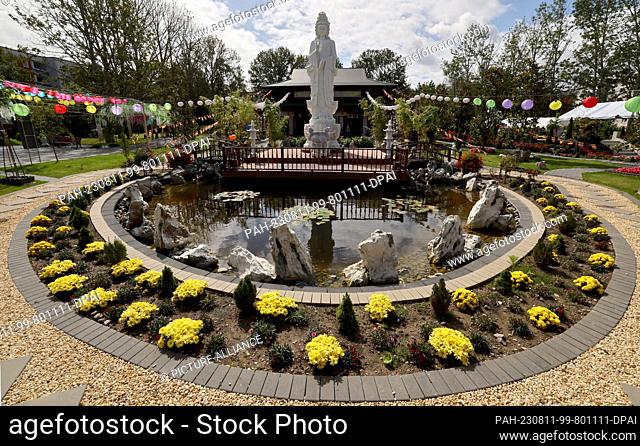 PRODUCTION - 10 August 2023, Mecklenburg-Western Pomerania, Rostock-Lichtenhagen: A pond surrounded by a flower bed and a sculpture stand in front of the Loc...