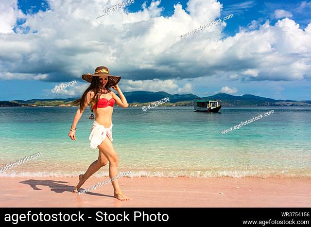 Full length of a happy young woman wearing straw hat and a trendy swimwear while walking through shallow sea water on the beach in Komodo Island, Indonesia