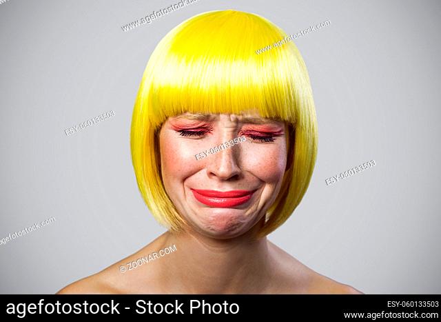 Portrait of sad alone cute young woman with freckles, red makeup and yellow wig, crying and depressed. indoor studio shot, isolated on gray background