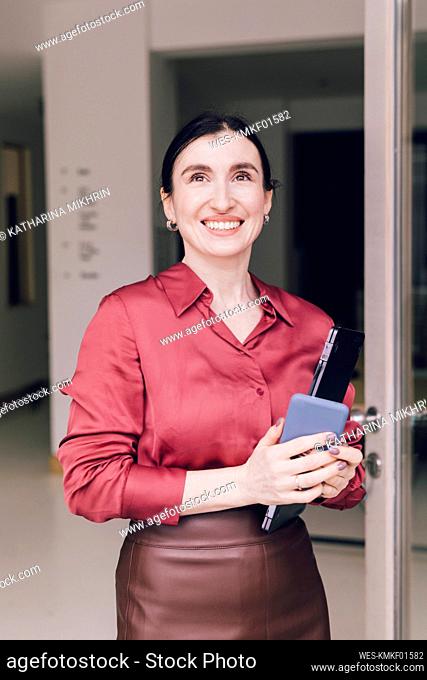 Smiling female entrepreneur with mobile phone and laptop standing at door in home office