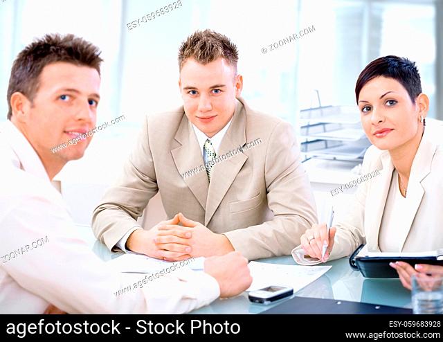 Businessteam of three working together, sitting around a desk, looking at camera