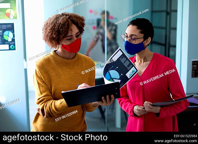 Two mixed race women wearing masks talking in an office