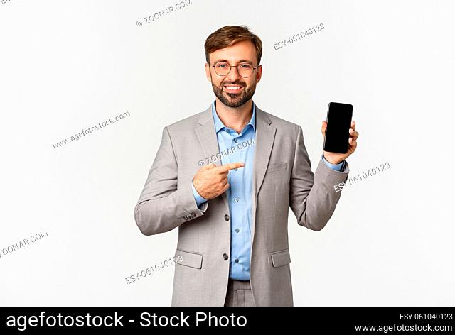 Handsome smiling businessman with beard, wearing grey suit and glasses, pointing finger at mobile phone screen, showing app, standing over white background