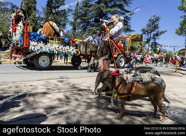 14 October 2023, Argentina, Villa General Belgrano: A photographer in lederhosen and with dogs on a leash takes pictures of the Oktoberfest parade