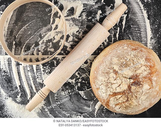 round baked bread, rolling pin, sieve and white wheat flour scattered on a black table, top view