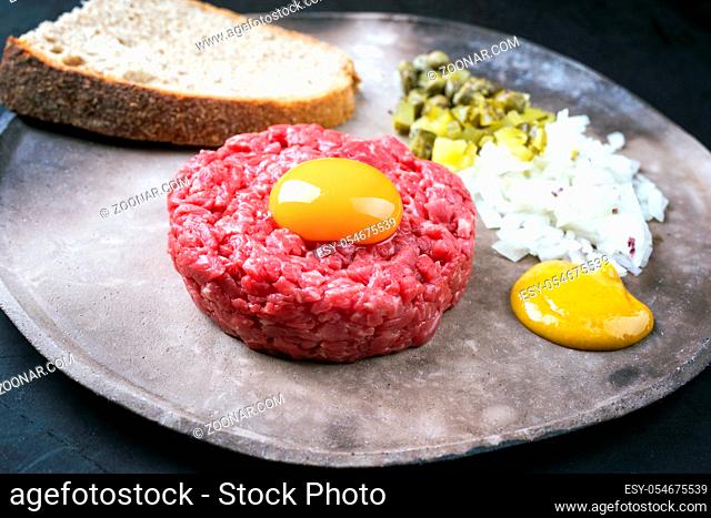 Gourmet tartar raw from beef fillet with yellow of the egg, gherkin and farmhouse bread as closeup on a modern design rustic plate