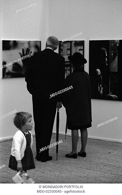 An elderly couple and a little girl visiting the Biennial fair dedicated to photography. Cologno Monzese, 1966