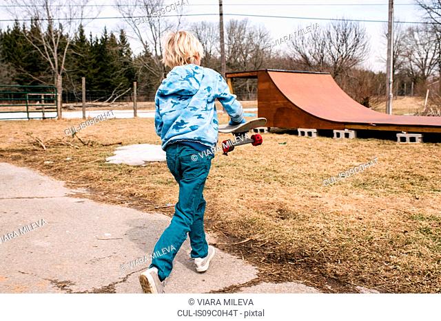 Blond boy carrying skateboard toward rural skateboard ramp, rear view