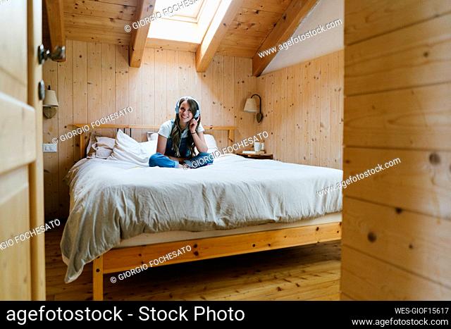 Smiling young woman wearing wireless headphones sitting on bed