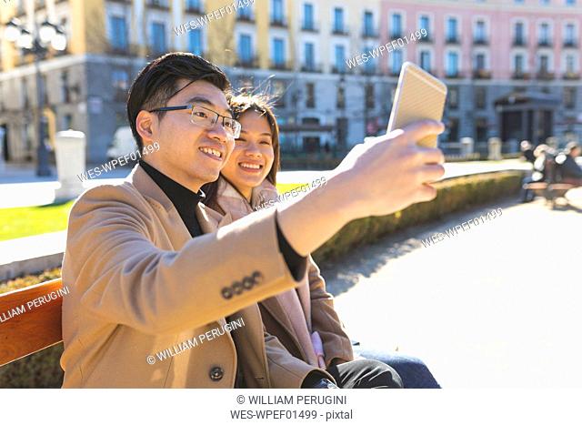 Spain, Madrid, young couple resting on a bench and taking a selfie in the city